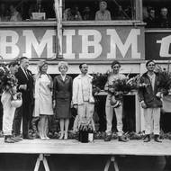 Henry Ford II s řidiči Fordu po jejich úspěchu ve Francii, 19. června 1966. Na stupních vítězů jsou vítězové závodu Bruce McLaren (vlevo) a Chris Amon (zcela vpravo) s Jo Siffert a Colin Davis, vítězové kategorie sportovních prototypů do objemu 2000. Švýcar Jo Siffert drží v ruce přilbu s typickým designem helvétskeho kříže.