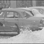 Muž odklízí sníh z parkovacího stání pro vozy Volha patřící tehdejší taxislužbě. 