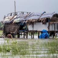 Muž po pás ve vodě řeky Shibsa opravuje rybářské sítě, zatímco jeho žena vaří v jejich domě na kůlech ve vesnici Jhulonto Para. (13. července)