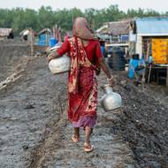 Champa Mondal, vysídlená kvůli změně klimatu, kráčí s nádobami s pitnou vodou po břehu řeky Shibsa. (12. července)
