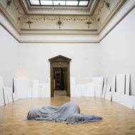 Uprostřed leží obryně zakrytá šedivou dekou a nazvaná S_ní, po obvodu místnosti je instalace Všechna včera.