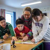 Osada rozprostírající se pod kopcem se může pochlubit školou, obchodem s potravinami a servisním domem, kde se mohou obyvatelé sprchovat a prát prádlo.
(Dánská učitelka Vanilla Mathiassen s dětmi ve škole v Kapisillitu, 20. ledna.)